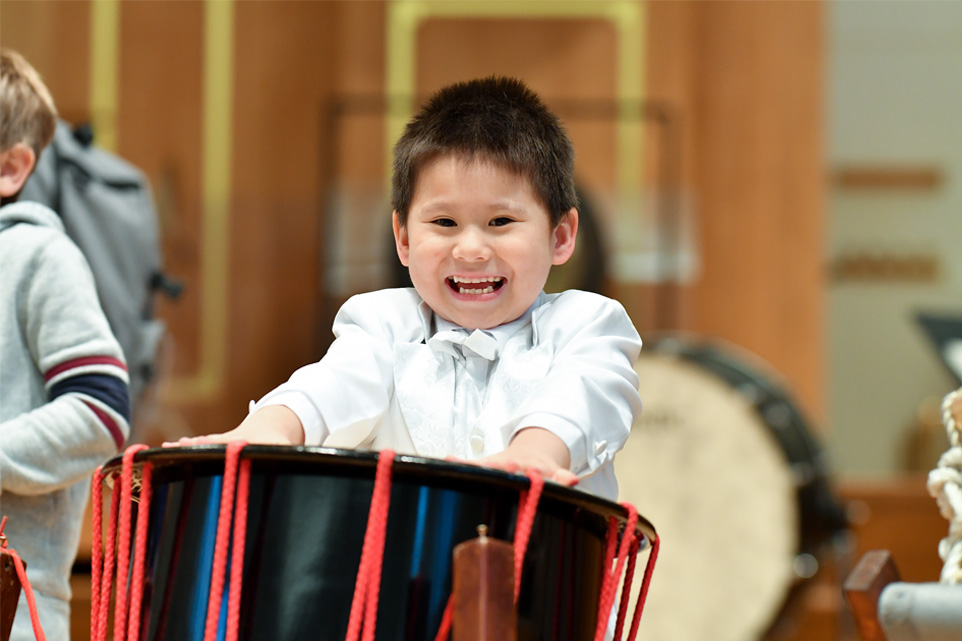 A young boy, wearing a white shirt, performing on a drum, smiling at the camera.